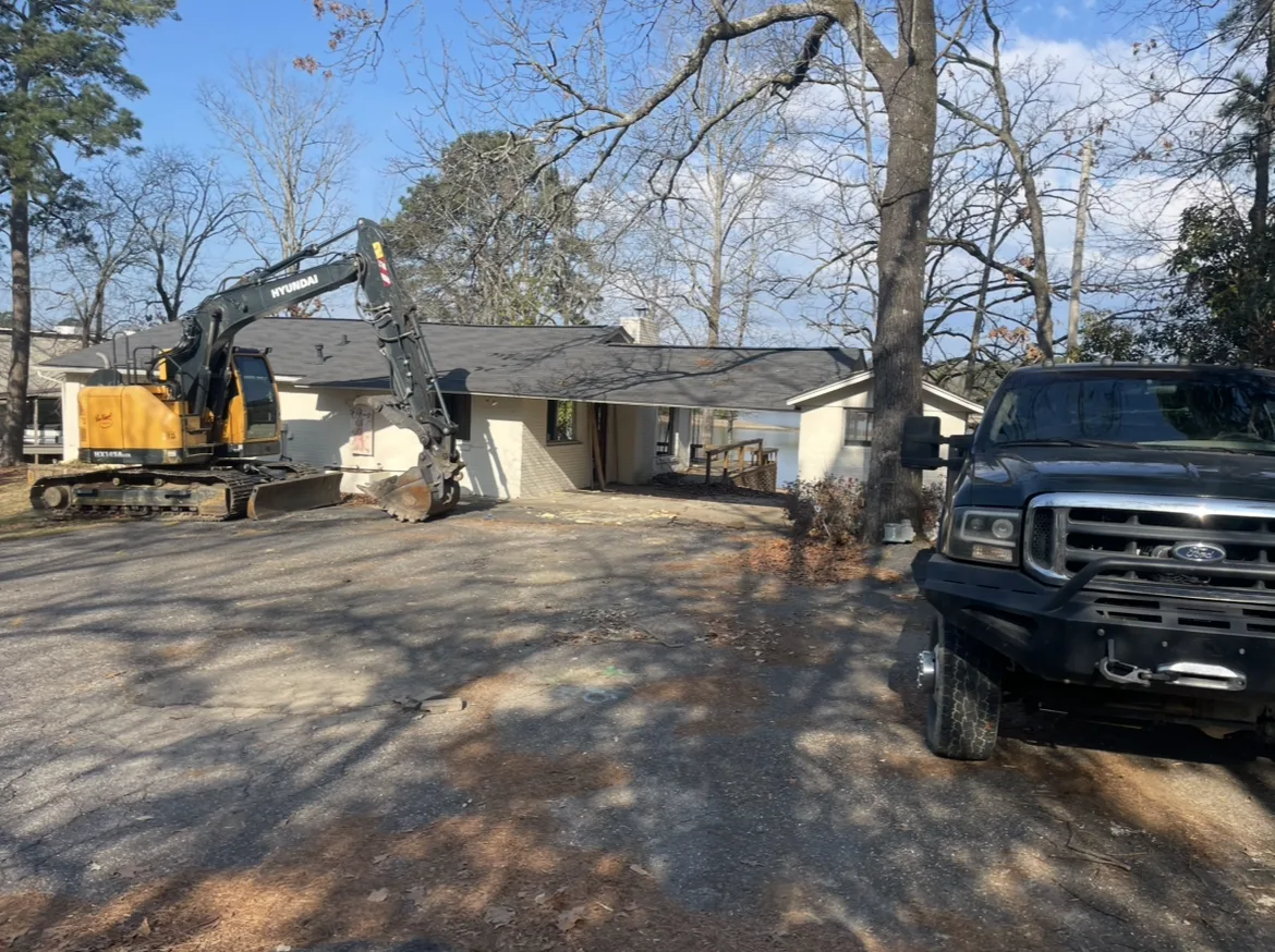 Tracked excavator staged beside a single-story brick house before demolition on a Hot Springs area job, pickup truck in the foreground